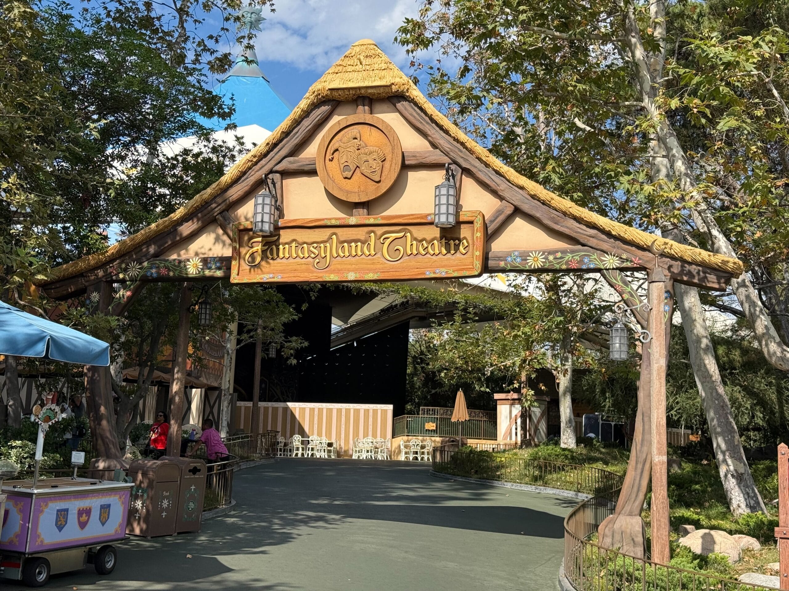Wooden entrance sign reading "Fantasyland Theatre" at Disneyland, surrounded by trees, lanterns, and a stage in the background.