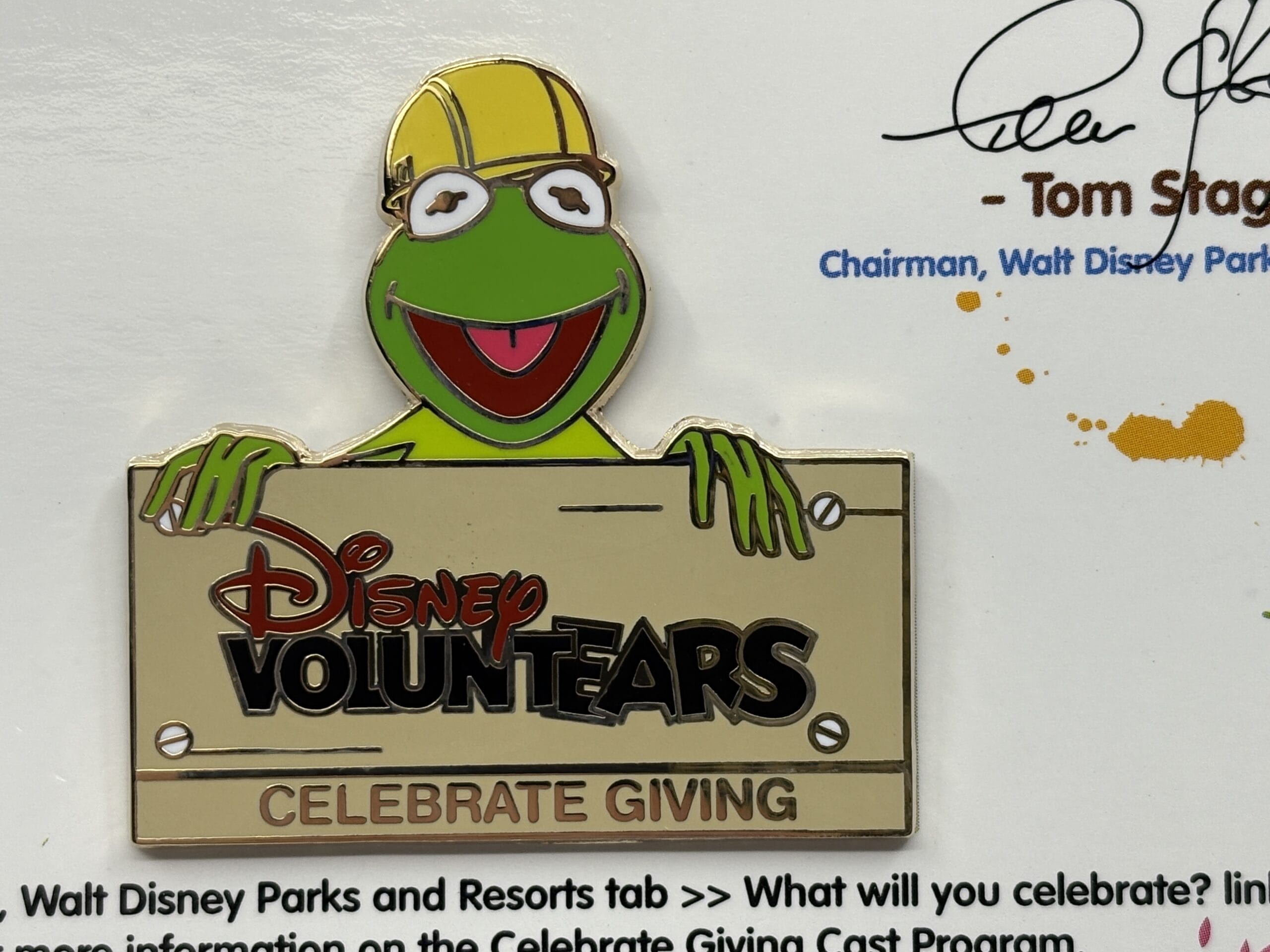 Kermit the Frog in a yellow hard hat holds a "Disney VoluntEARS Celebrate Giving" sign at the WDWNT Summer Auction.