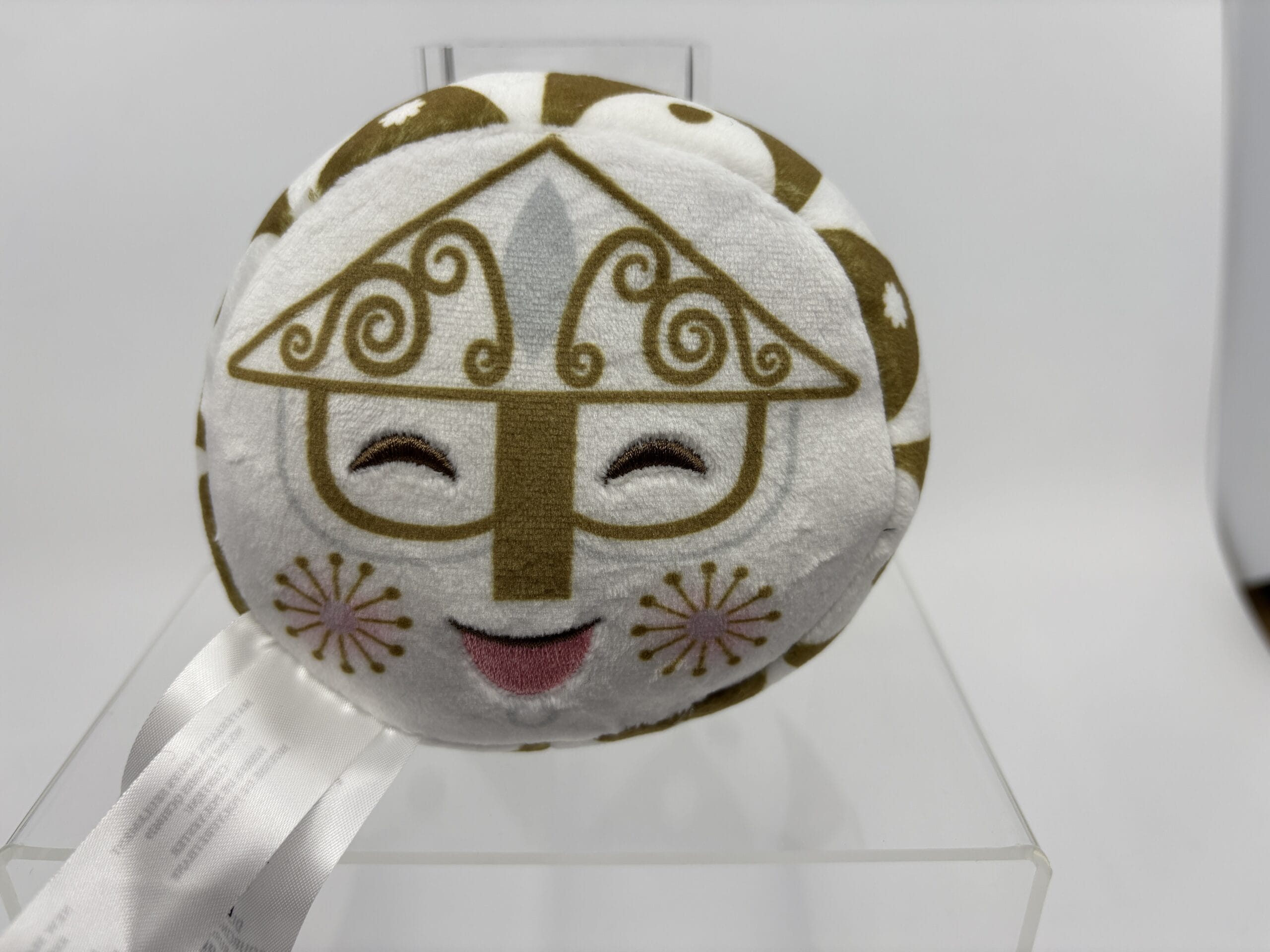 Round plush toy with a smiling face, gold patterns, and pink mouth, featured in the WDWNT summer auction on a clear stand.