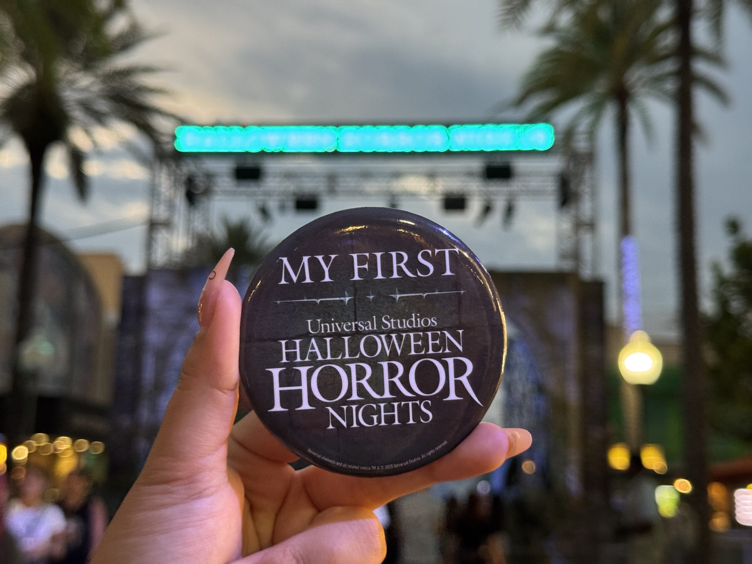 Hand holding a "My First Universal Studios Halloween Horror Night 2025" button with palm trees and stage in the background.