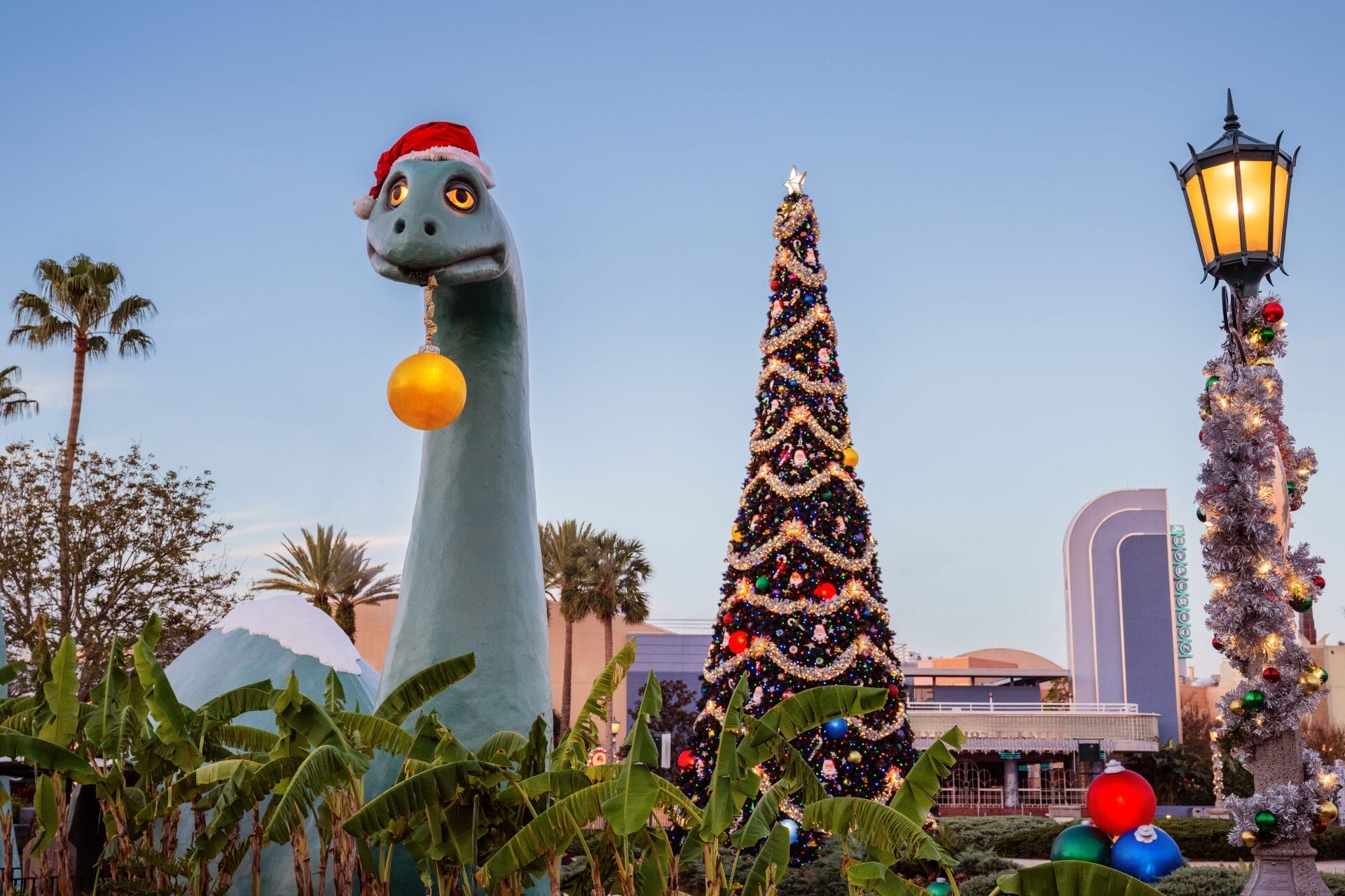A dinosaur statue with a Santa hat and ornament stars in this festive Daily Recap by a decorated tree and holiday lights.
