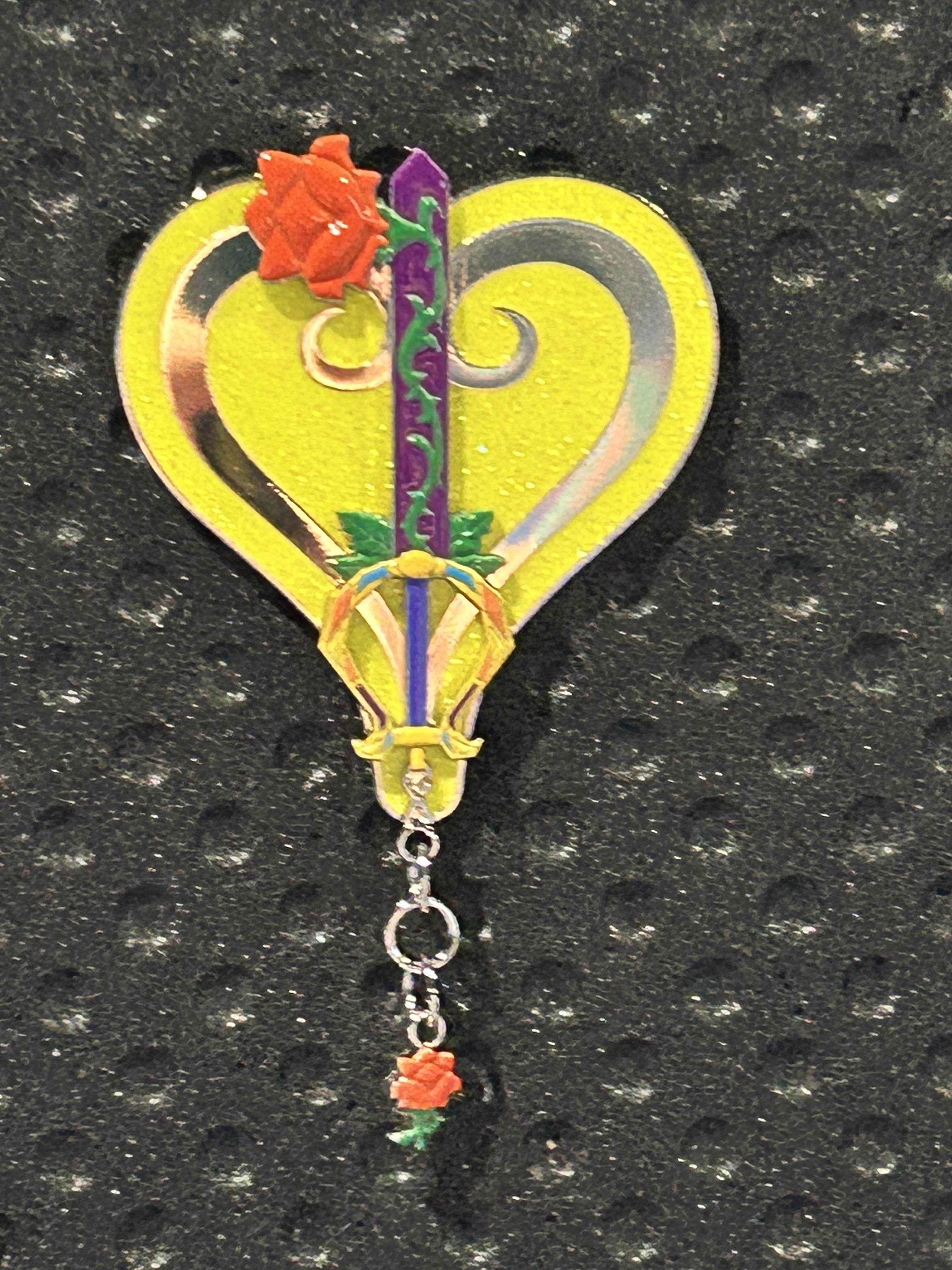 A heart-shaped yellow Kingdom Hearts pin with sword and rose, Pin of the Month 2026, on a textured black background.