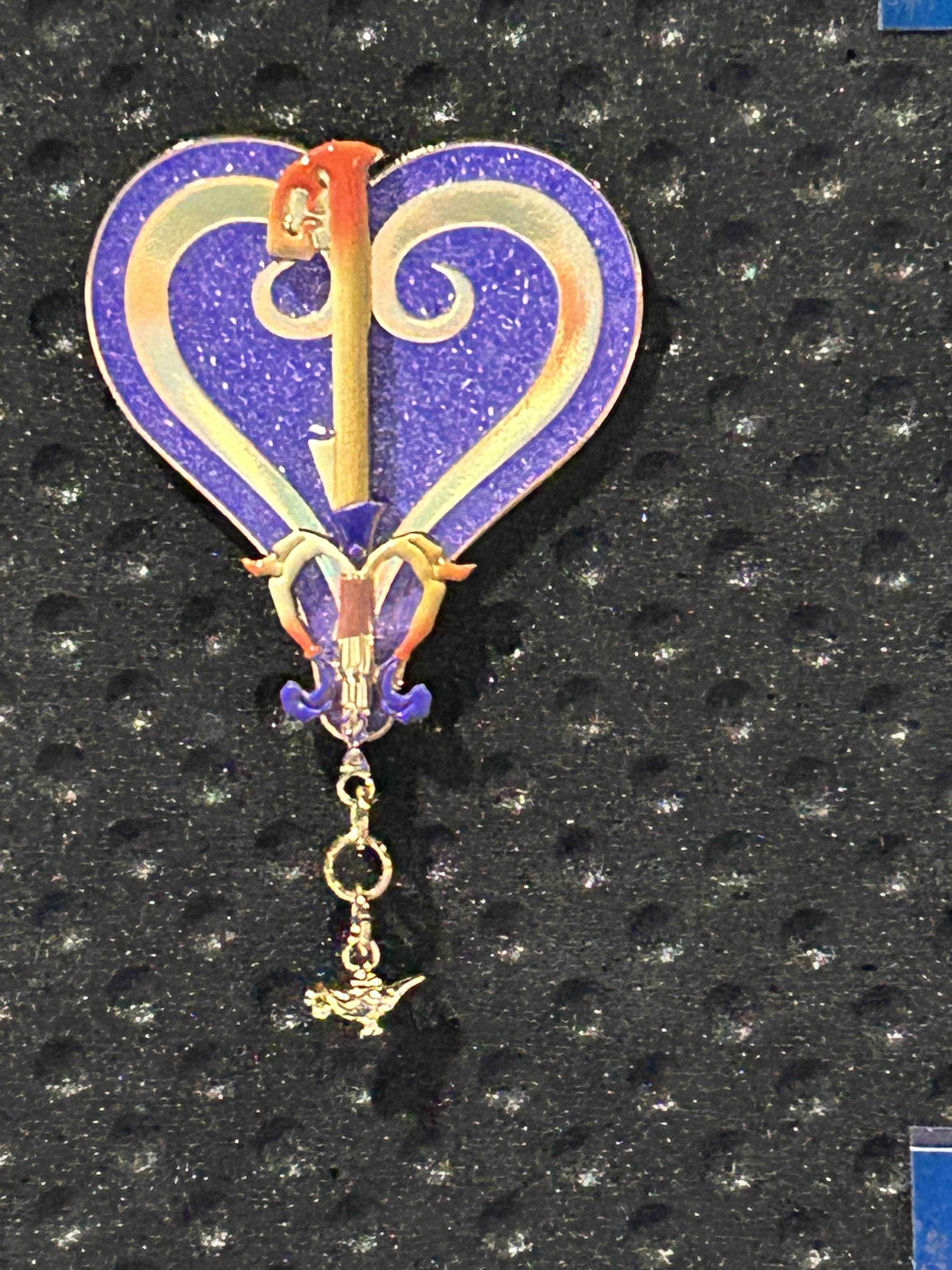 A purple heart-shaped Pin of the Month with gold details and a dangling charm, inspired by Kingdom Hearts.