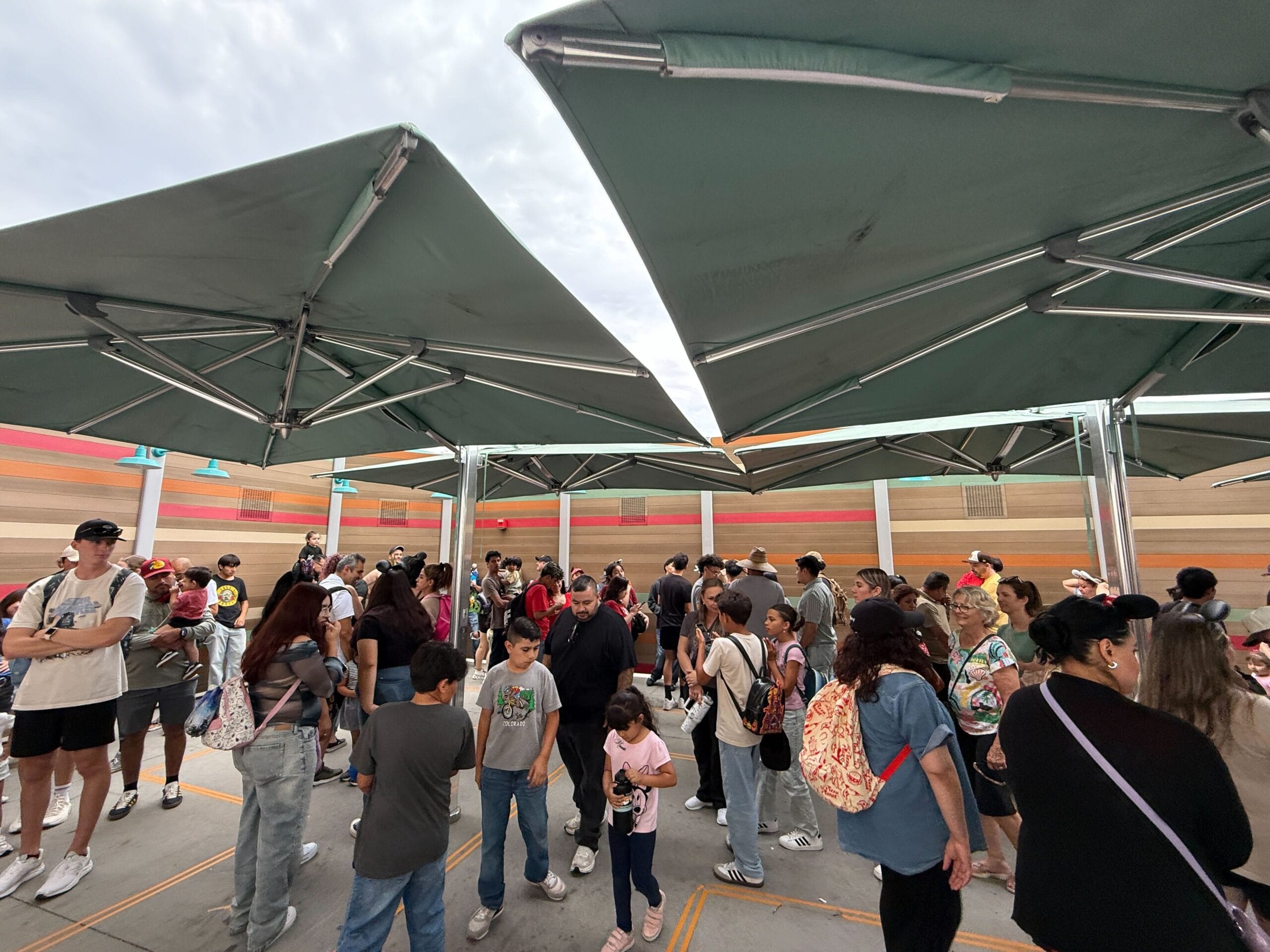People wait in an outdoor queue under green umbrellas at a venue with striped walls and a cloudy sky above.