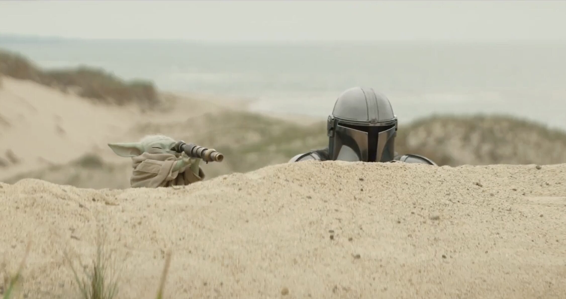 The titular characters of Grogu and the Mandalorian hide behind a sand dune as Grogu uses a spyglass in a scene from the upcoming movie.
