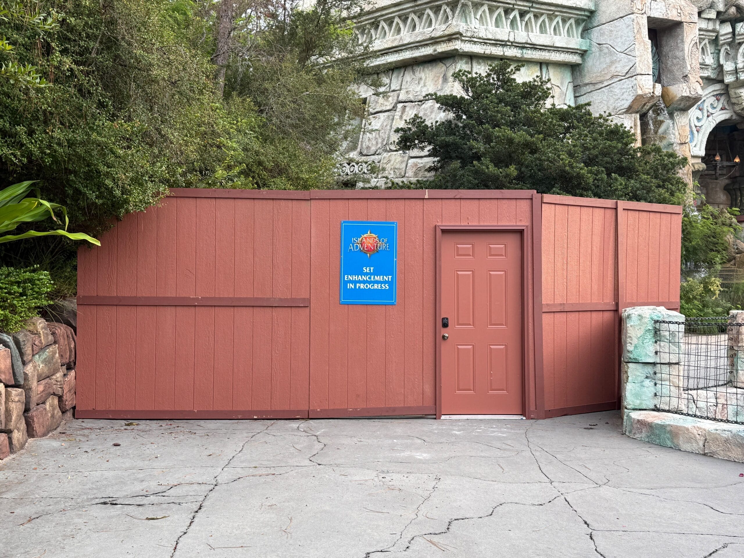 Brown construction wall with a "17+ enhancement in progress" sign, featured in today's Daily Recap near themed building and greenery.