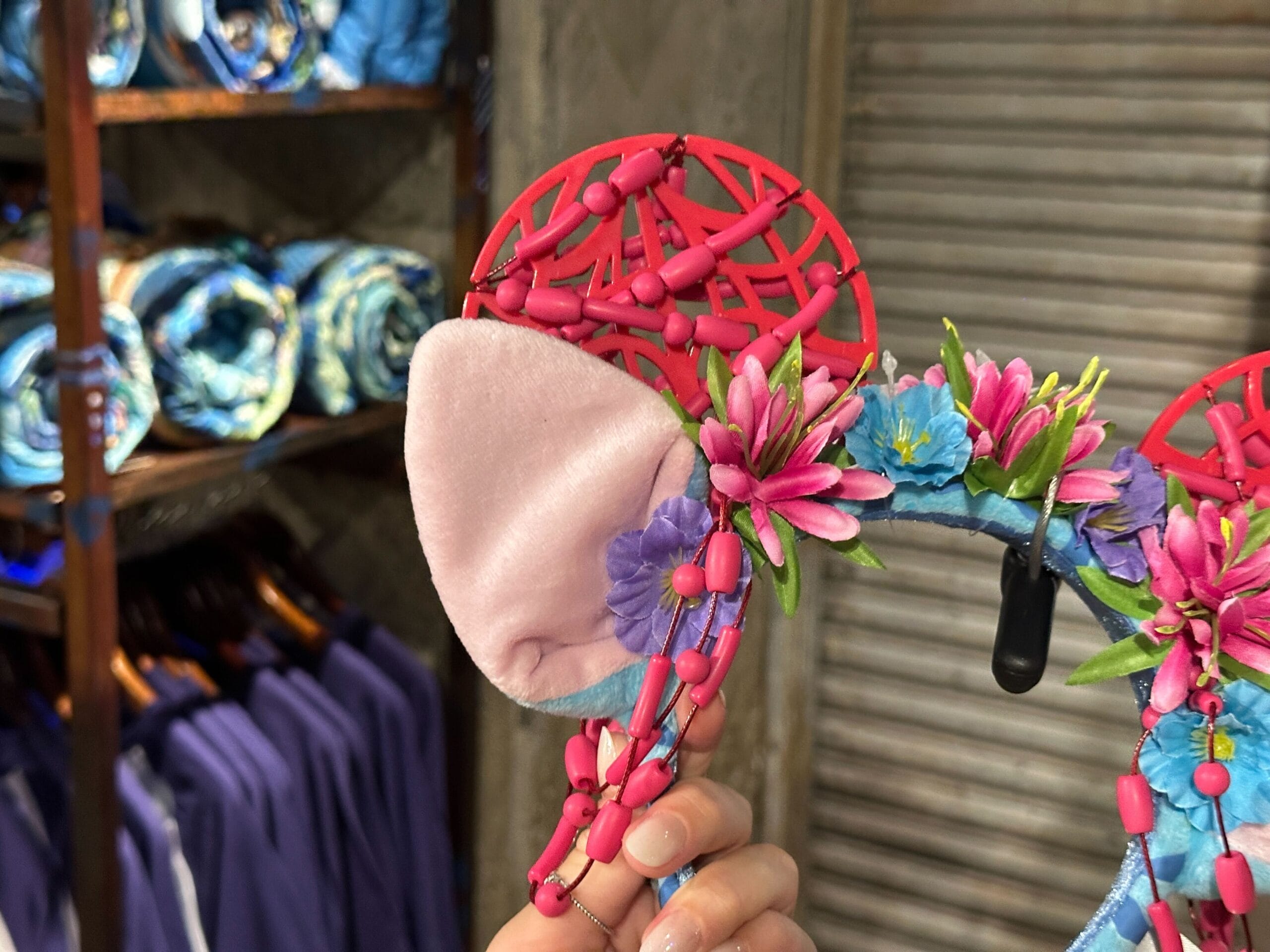 A hand holds a colorful floral mouse ear headband in a store, with blue clothing displayed in the background.