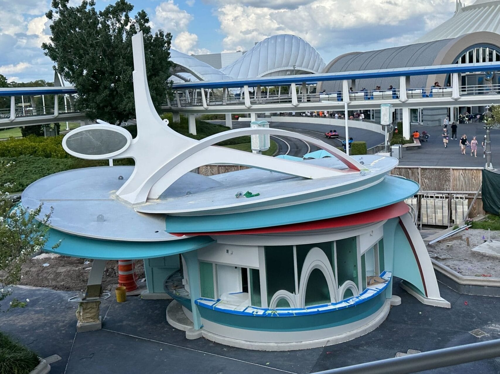 Futuristic, closed kiosk with white arches near Magic Kingdom's walkway and domed buildings under a cloudy blue sky.