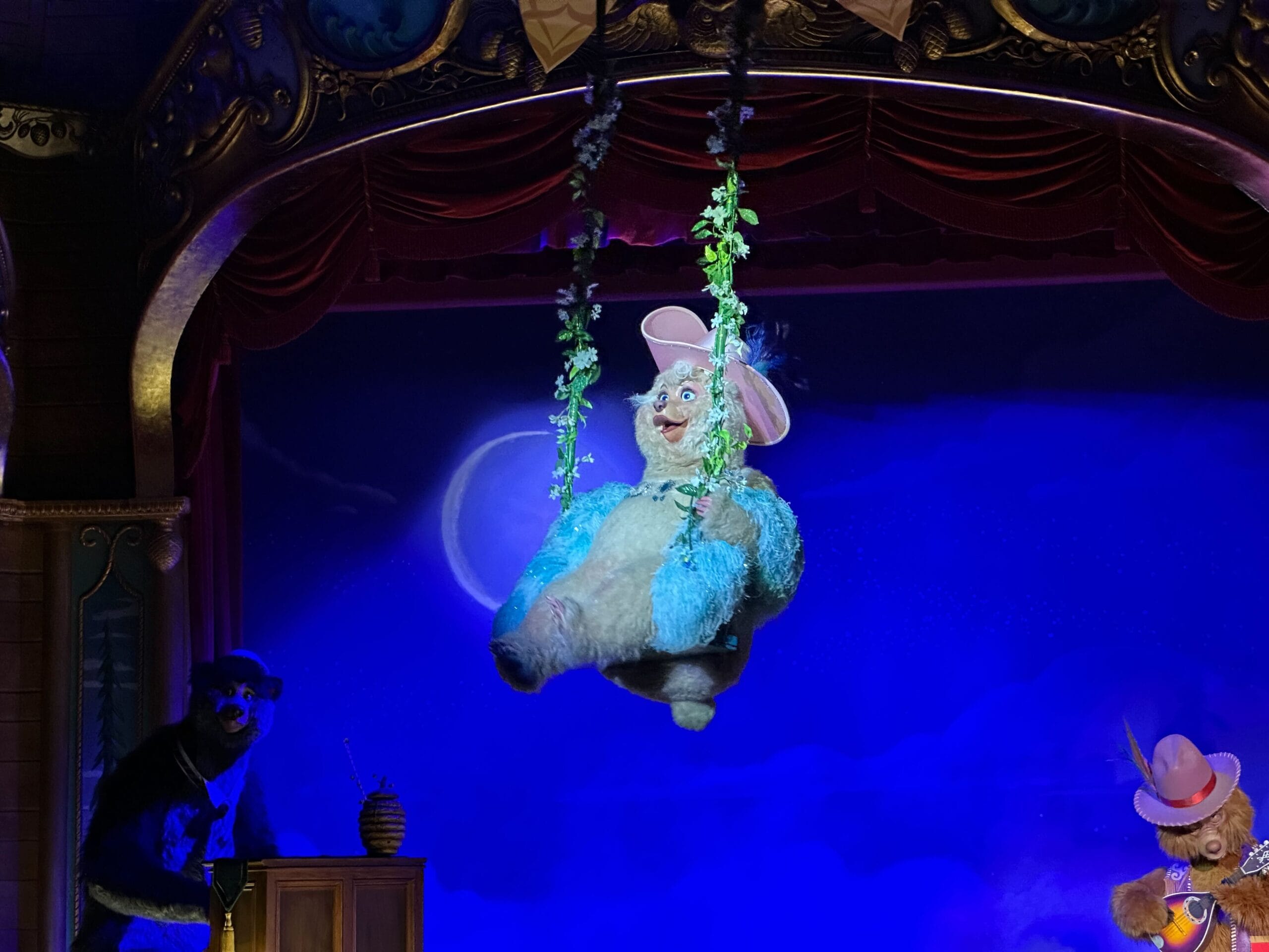 Teddi Barra, the blue bear puppet, swings on a vine in the Country Bear Musical Jamboree under stage lights and red curtains.