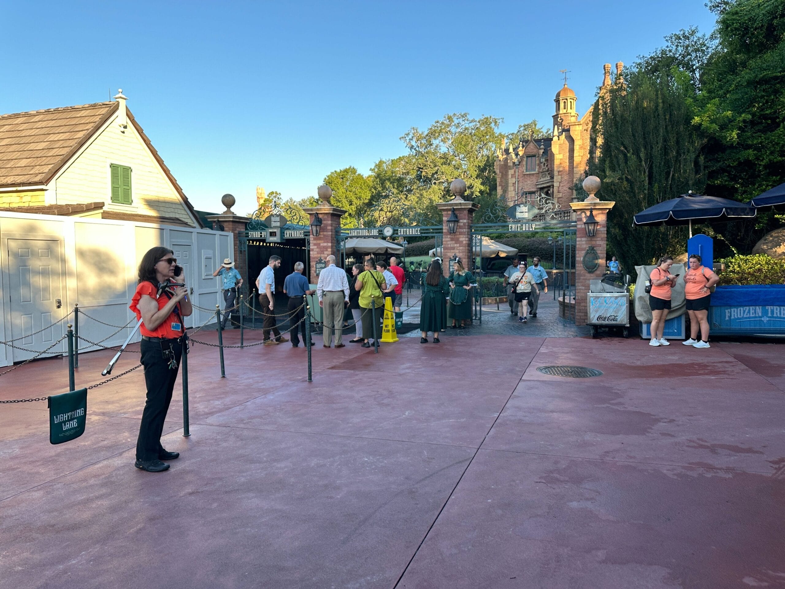 A woman with a microphone stands near a gated park entrance as people wait in line by the Haunted Mansion attraction.